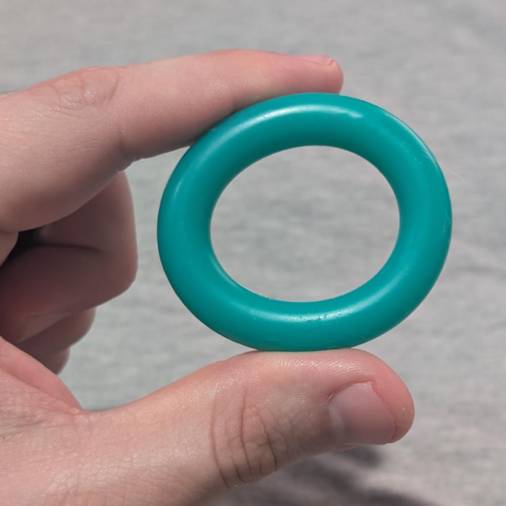 A hand holding a plain teal silicone cock ring between thumb and forefinger against a pale grey background.